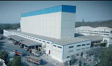 A large, modern industrial manufacturing facility with a blue and white exterior and commercial trucks docked at the loading bays.