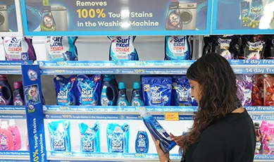 A customer in a supermarket aisle examining a bottle of Surf Excel laundry detergent from a fully stocked shelf.