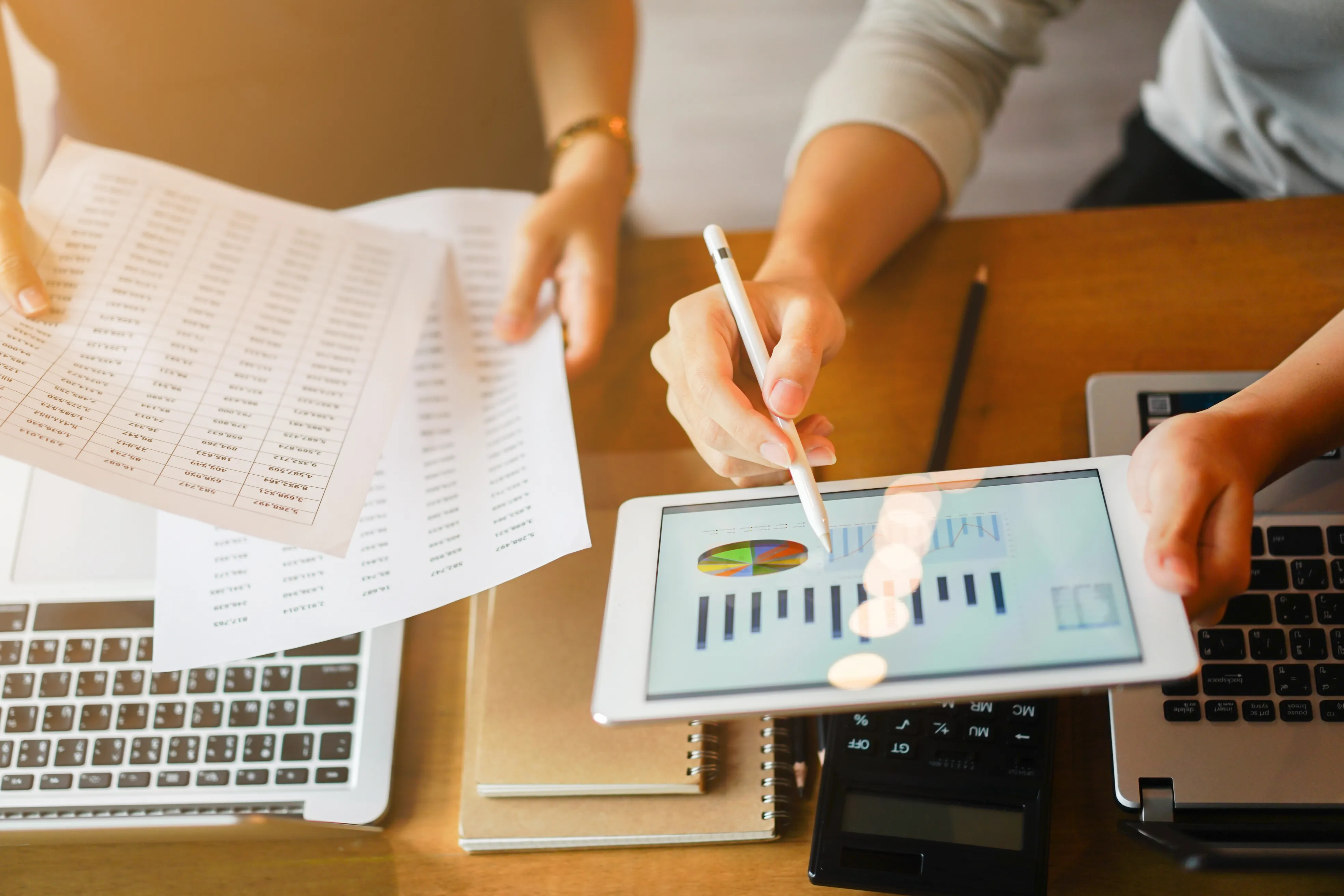 Two professionals collaborating at a desk, analyzing business performance metrics across printed spreadsheets and a digital tablet.